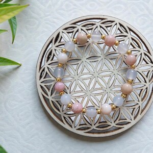 Peut inclure: Un bracelet de perles alternant des pierres précieuses roses, blanches et pêche, entrecoupées de petites perles dorées. Le bracelet est présenté sur un dessous de verre rond en bois avec un motif géométrique. Le fond est blanc.