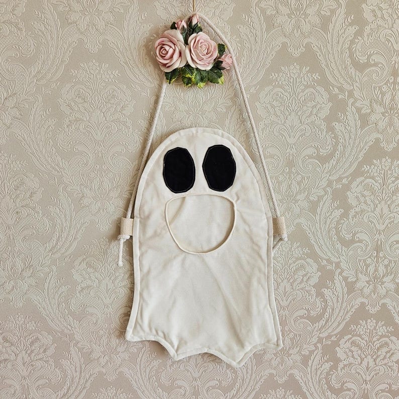 Może przedstawiać: Biała dekoracja na Halloween w kształcie ducha z czarnymi owalnymi oczami i otworem na usta. Wisi na sznurku przymocowanym do dekoracyjnej kompozycji kwiatowej z r&oacute;żowych r&oacute;ż i zielonych liści.