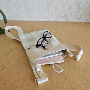 Op de afbeelding: Een beige en cr&egrave;me geruite crossbodytas met een cr&egrave;mekleurige band en goudkleurige hardware. De tas bevat een roze notitieboek, een wit notitieboek en een bril met zwart montuur. Een kleine gouden ring ligt op de tas.