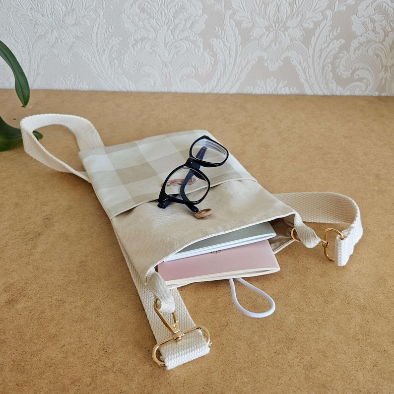 Op de afbeelding: Een beige en cr&egrave;me geruite crossbodytas met een cr&egrave;mekleurige band en goudkleurige hardware. De tas bevat een roze notitieboek, een wit notitieboek en een bril met zwart montuur. Een kleine gouden ring ligt op de tas.