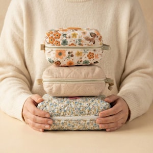May include: Three stacked, rectangular, quilted cosmetic bags. The top bag features a floral pattern with orange, yellow, and green flowers on a white background. The middle bag is light pink with a quilted design. The bottom bag has a blue floral pattern.