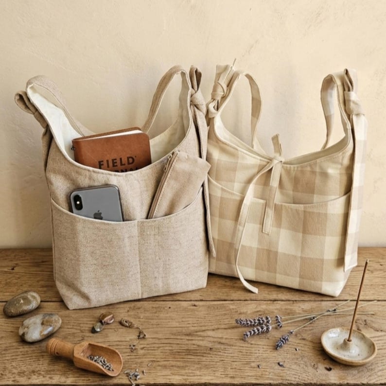 May include: Two beige tote bags with shoulder straps sit on a wooden surface. One bag has a pocket with a phone, notebook, and a small pouch. The other bag has a plaid pattern. The word "FIELD" is visible on the notebook.