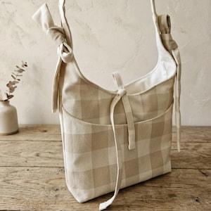 May include: A beige and white checkered shoulder bag with a front pocket and knotted strap details. The bag has a soft, casual design and is made of a woven fabric. The bag is sitting on a wooden surface.