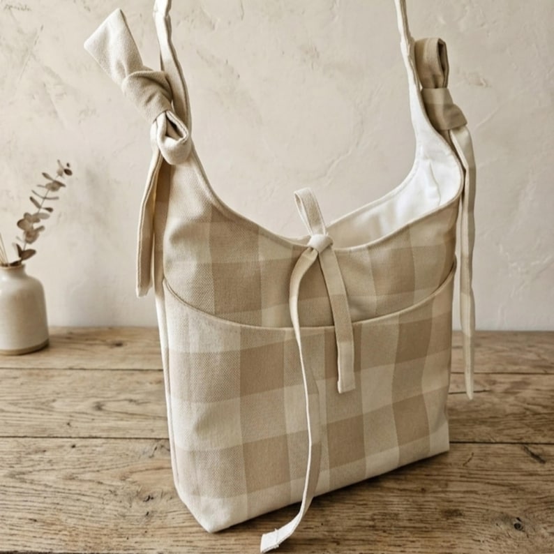 May include: A beige and white checkered shoulder bag with a front pocket and knotted strap details. The bag has a soft, casual design and is made of a woven fabric. The bag is sitting on a wooden surface.
