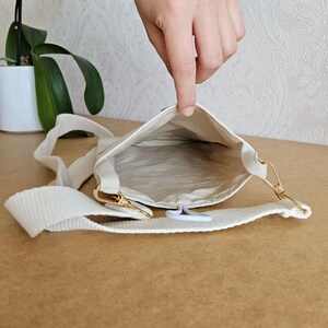 Op de afbeelding: Cr&egrave;mekleurige crossbodytas met een lange, verstelbare riem en goudkleurige hardware. De binnenkant is lichtbeige. De tas is open en onthult de binnenkant. De riem is gemaakt van geweven materiaal.