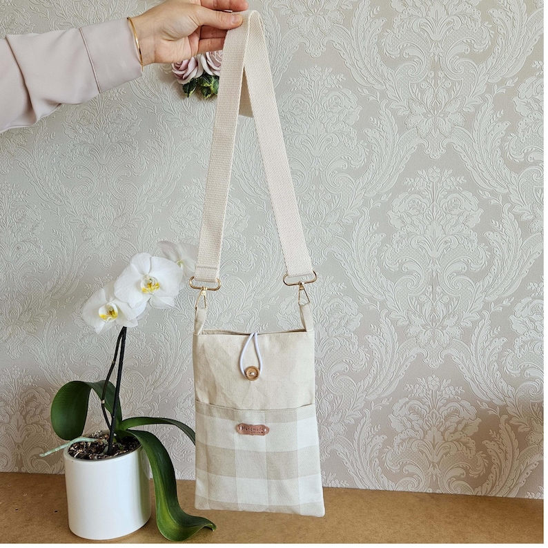 Op de afbeelding: Een beige en witte crossbody tas met een ruitpatroon op de onderste helft. De tas heeft een lange, verstelbare riem en goudkleurige hardware. Een kleine, ronde knoop sluit de bovenklep af. De tas wordt door een hand omhoog gehouden.