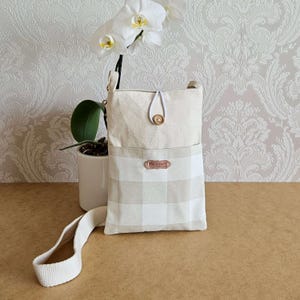 Op de afbeelding: Een beige crossbody tas met een klepsluiting en een lichtbeige en wit geruit vak. De tas heeft een lange, verstelbare riem en een houten knoopsluiting. Handgemaakt met een klein "Handmade" label.