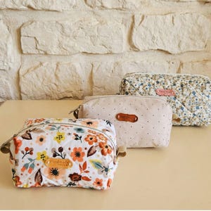 May include: Three handmade cosmetic bags with floral and quilted patterns. The bags feature a variety of colours, including white, orange, and blue. Each bag has a zip closure and a small "Handmade" label.