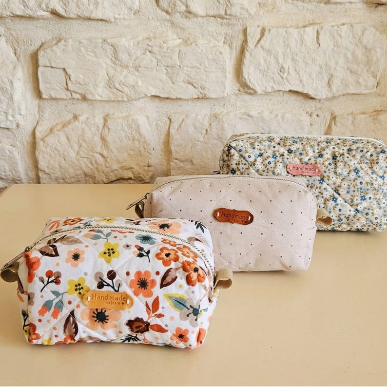 May include: Three handmade cosmetic bags with floral and quilted patterns. The bags feature a variety of colours, including white, orange, and blue. Each bag has a zip closure and a small "Handmade" label.
