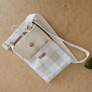 Op de afbeelding: Een beige crossbodytas met een lichtbeige en wit geblokt patroon op de voorklep. De tas heeft een beige interieur, een verstelbare beige riem en goudkleurige hardware. Een klein, bruin label leest "hand made".