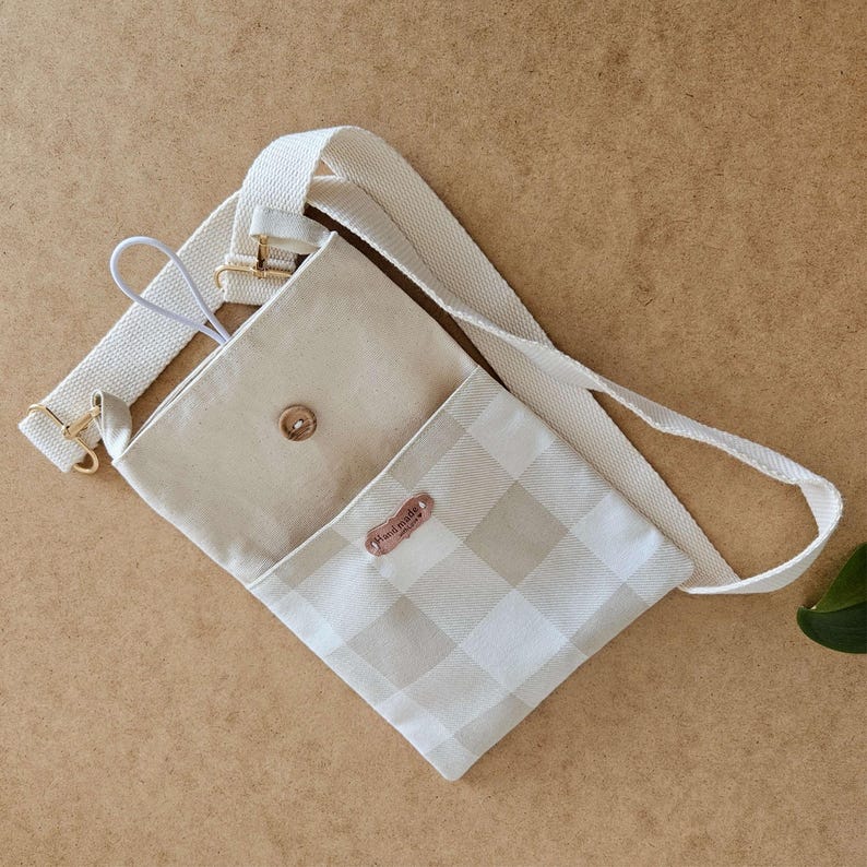 Op de afbeelding: Een beige crossbodytas met een lichtbeige en wit geblokt patroon op de voorklep. De tas heeft een beige interieur, een verstelbare beige riem en goudkleurige hardware. Een klein, bruin label leest "hand made".