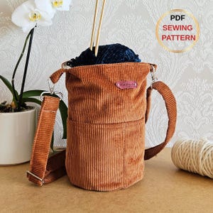 May include: A brown corduroy yarn bag with a shoulder strap and handle, holding blue yarn and knitting needles. The bag has a small "Handmade" label. The image also includes a "PDF Sewing Pattern" graphic. The bag is designed for knitting.