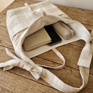 May include: A beige and white checkered tote bag with a long strap, open to reveal a journal, a smartphone, and a pencil case. The bag is made of a soft fabric and sits on a wooden surface.
