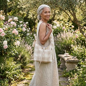 May include: A beige and white checkered shoulder bag with a matching strap is carried by a person in a garden. The bag has a rectangular shape with two pockets on the front. The person is wearing a beige dress. The background features a stone bench and blooming flowers.
