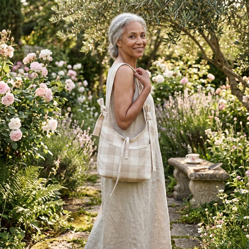 May include: A beige and white checkered shoulder bag with a matching strap is carried by a person in a garden. The bag has a rectangular shape with two pockets on the front. The person is wearing a beige dress. The background features a stone bench and blooming flowers.