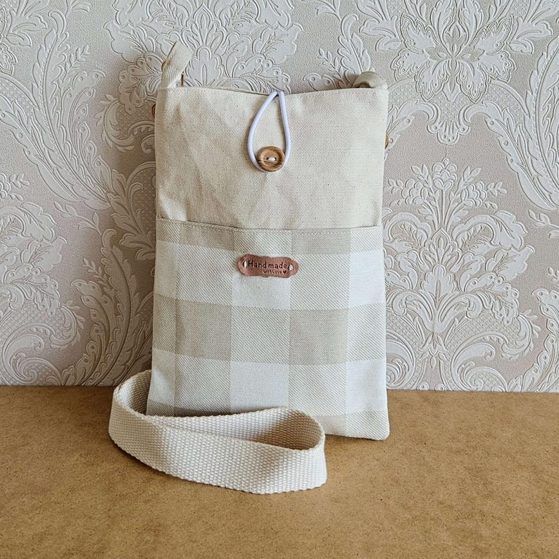 Op de afbeelding: Een beige en witte crossbody tas met een geruit patroon op de voorzak. De tas heeft een lange, verstelbare riem en een houten knoopsluiting. Een klein label leest "Handmade with love".