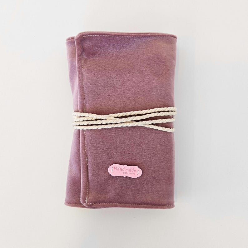 May include: A dusky pink velvet roll-up case secured with a cream rope. A small pink label with the words "Handmade with Love" is attached. The case is designed to hold items such as makeup brushes or art supplies. The case is approximately 20 cm in length.