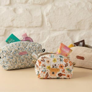 May include: Three small, quilted cosmetic bags with floral patterns. One bag is light blue with small white and blue flowers, another has a white background with orange and yellow flowers, and the third is beige with small dots. Each bag is open and filled with toiletries.
