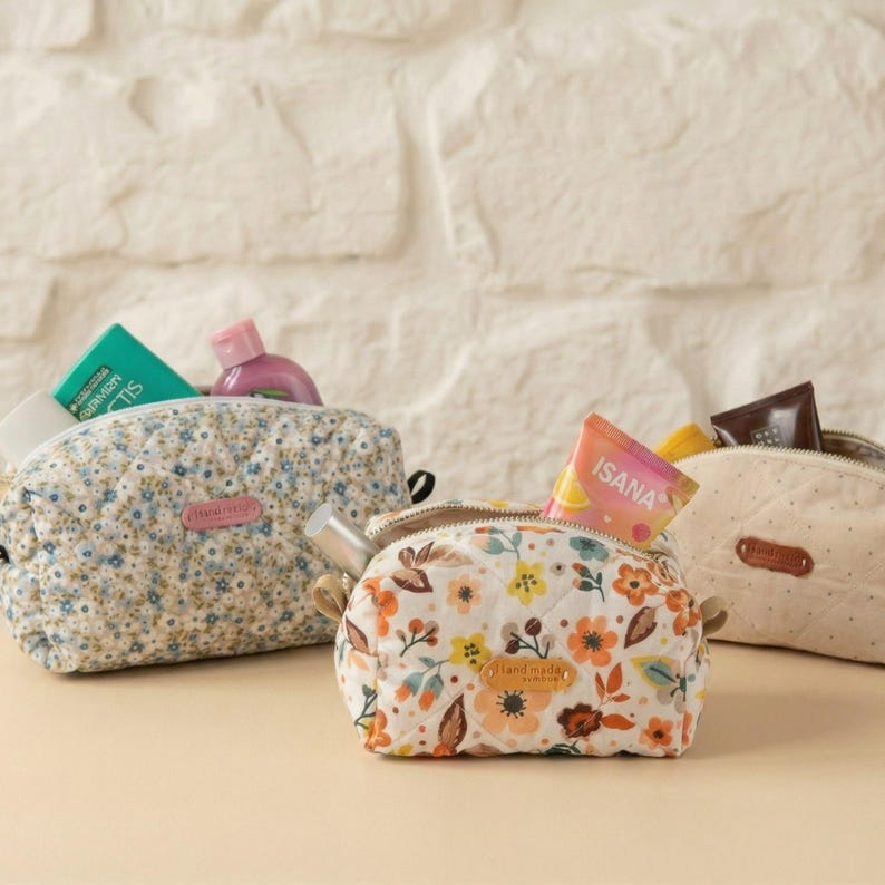 May include: Three small, quilted cosmetic bags with floral patterns. One bag is light blue with small white and blue flowers, another has a white background with orange and yellow flowers, and the third is beige with small dots. Each bag is open and filled with toiletries.
