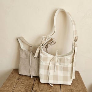 May include: Two shoulder bags in neutral tones. One is solid beige, the other has a beige and white checkered pattern. Both have a front pocket and a drawstring closure. The bags are made of a soft fabric and have long straps.