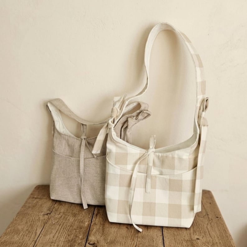 May include: Two shoulder bags in neutral tones. One is solid beige, the other has a beige and white checkered pattern. Both have a front pocket and a drawstring closure. The bags are made of a soft fabric and have long straps.