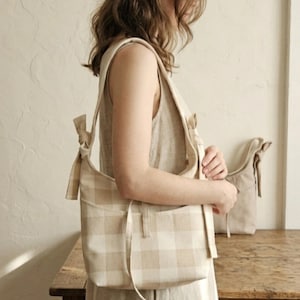 May include: A beige and white plaid shoulder bag with a matching tote bag in the background. The shoulder bag has a long strap and a pocket on the front. The bags are made of a soft, woven fabric.