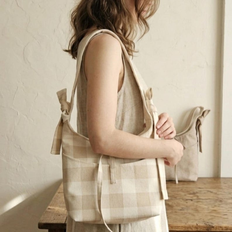 May include: A beige and white plaid shoulder bag with a matching tote bag in the background. The shoulder bag has a long strap and a pocket on the front. The bags are made of a soft, woven fabric.