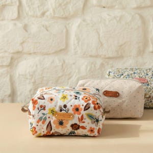 May include: Three small, quilted cosmetic bags with floral patterns and the word "Handmade" on a leather tag. The bags feature a variety of colours, including orange, yellow, and blue, against a white background.