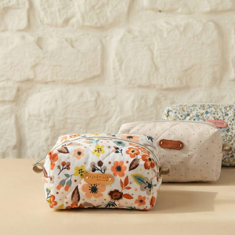 May include: Three small, quilted cosmetic bags with floral patterns and the word "Handmade" on a leather tag. The bags feature a variety of colours, including orange, yellow, and blue, against a white background.