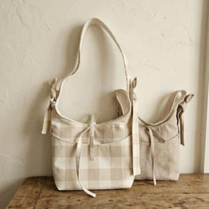 May include: Two beige shoulder bags. One bag features a tan and white checkered pattern, while the other is a solid beige. Both bags have long straps and decorative bows.