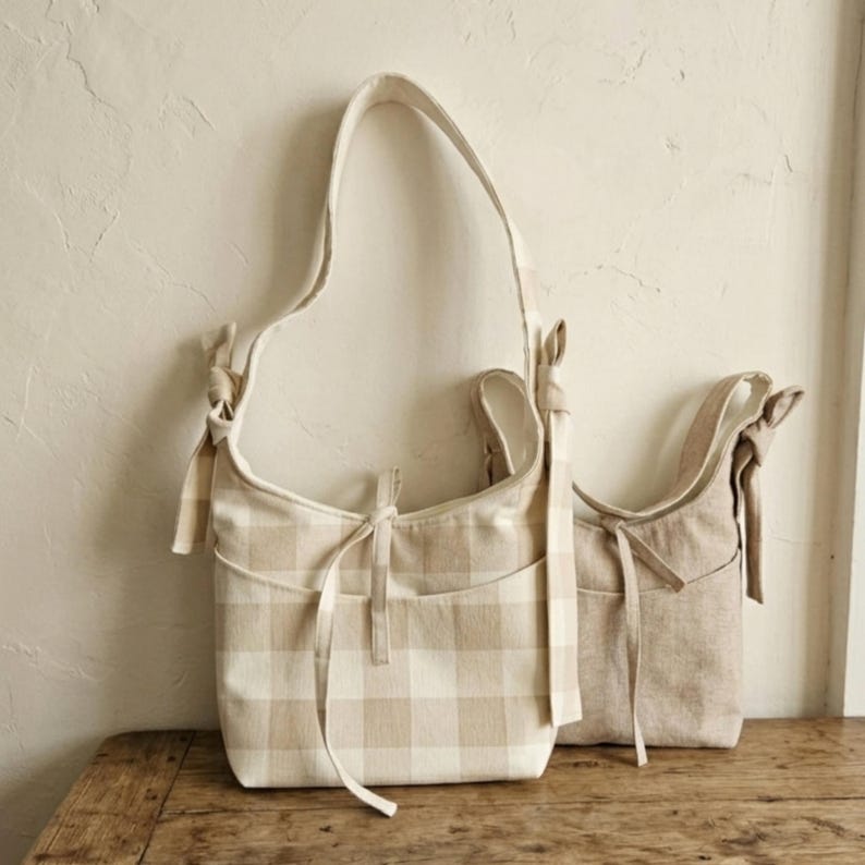 May include: Two beige shoulder bags. One bag features a tan and white checkered pattern, while the other is a solid beige. Both bags have long straps and decorative bows.