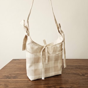 May include: A beige and white plaid shoulder bag with a front pocket and a tie closure. The bag has long straps that are tied at the top. The bag is sitting on a wooden surface.