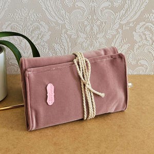 May include: A dusky pink velvet makeup brush roll secured with a cream rope. A small pink label with the text "handmade" is attached. The roll is closed and rests on a brown surface.