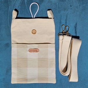 Op de afbeelding: Een handgemaakte stoffen tas in beige en cr&egrave;me met een ruitpatroon op de onderste helft. De tas heeft een houten knoopsluiting, een witte lus en een afneembare cr&egrave;mekleurige riem met goudkleurige sluitingen. Het label luidt "handmade with love".
