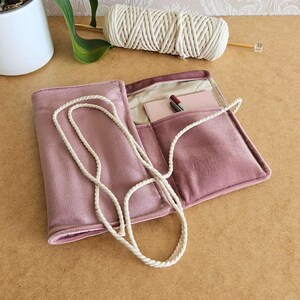 May include: A dusty rose-coloured, open, velvet-like case with a white rope strap. Inside, a small notepad and pen are visible. A spool of white yarn and a knitting needle are in the background. The case is on a light brown surface.