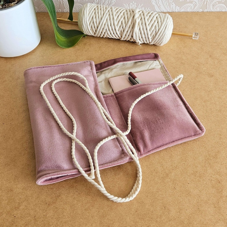 May include: A dusty rose-coloured, open, velvet-like case with a white rope strap. Inside, a small notepad and pen are visible. A spool of white yarn and a knitting needle are in the background. The case is on a light brown surface.
