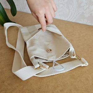 Op de afbeelding: Een beige crossbodytas met een geruite flap en een witte band. De tas heeft een goudkleurige sluiting en een knoopsluiting. De binnenkant is zichtbaar en een hand opent de flap.