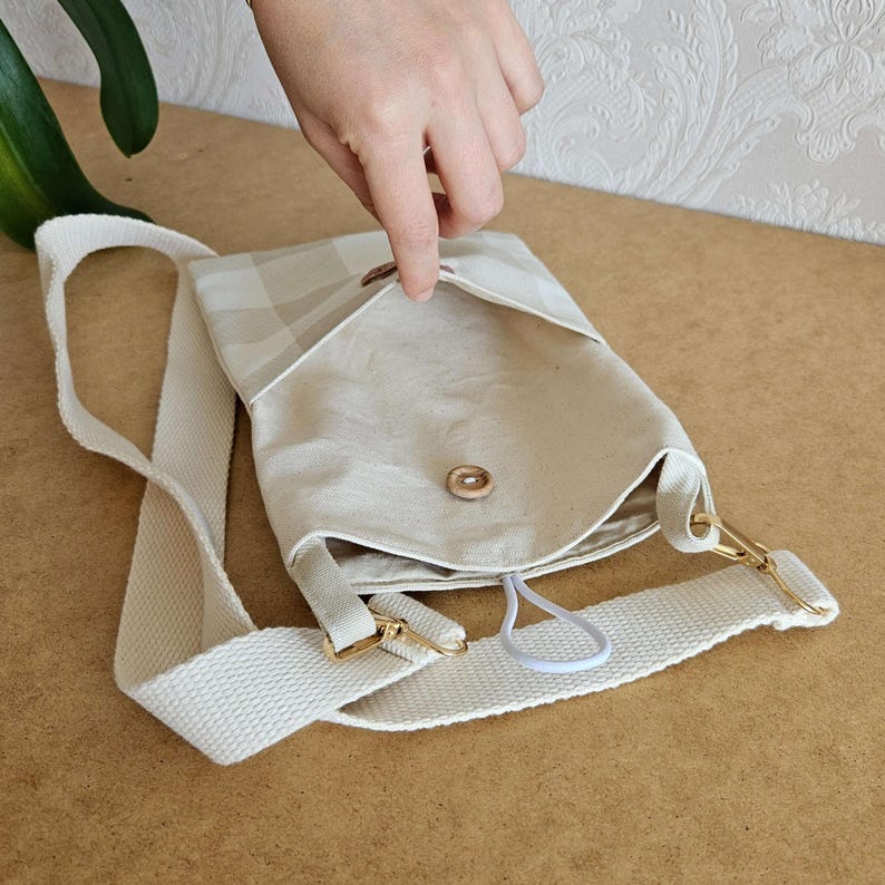 Op de afbeelding: Een beige crossbodytas met een geruite flap en een witte band. De tas heeft een goudkleurige sluiting en een knoopsluiting. De binnenkant is zichtbaar en een hand opent de flap.