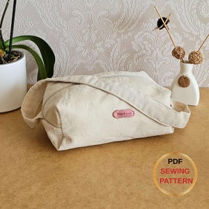Puede incluir: Un bolso de pana color crema con cierre de cremallera y correa para el hombro. El bolso tiene una etiqueta rosa "Handmade" y está sobre una superficie marrón. Un jarrón blanco con elementos decorativos está en el fondo. Un círculo dorado con el texto "PDF SEWING PATTERN" está en la esquina inferior derecha.