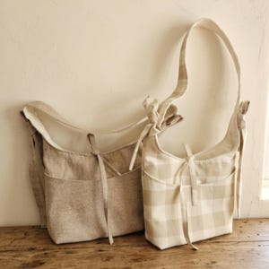 May include: Two beige shoulder bags with long straps. One bag is solid beige, and the other has a beige and white checkered pattern. Both bags have a pocket on the front and are tied with a bow.
