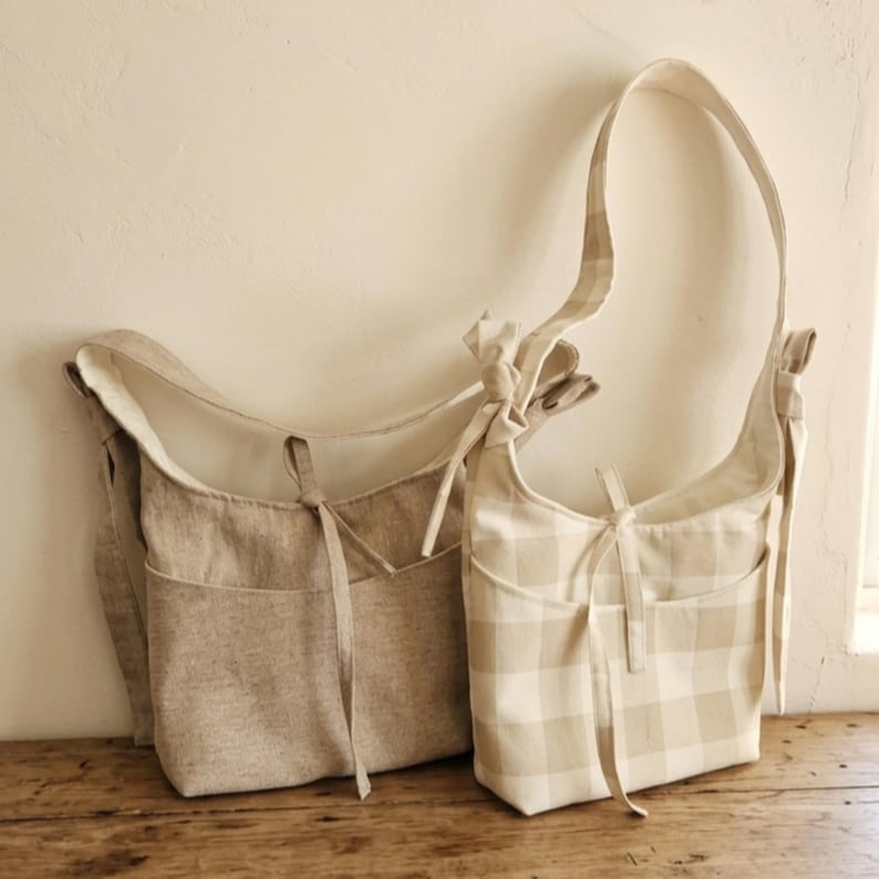 May include: Two beige shoulder bags with long straps. One bag is solid beige, and the other has a beige and white checkered pattern. Both bags have a pocket on the front and are tied with a bow.