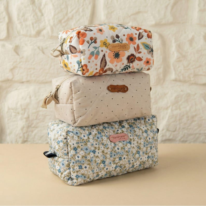 May include: Three quilted cosmetic bags stacked. The top bag features a floral pattern with orange, yellow, and brown flowers. The middle bag is beige with black dots. The bottom bag has a blue floral pattern. Each bag has a zipper.