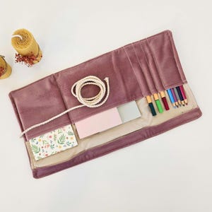 May include: A dusty rose velvet pencil roll, open to display its contents. It contains coloured pencils, notepads, and a floral card. A white rope is coiled on top. The roll is made of a soft, velvet material.
