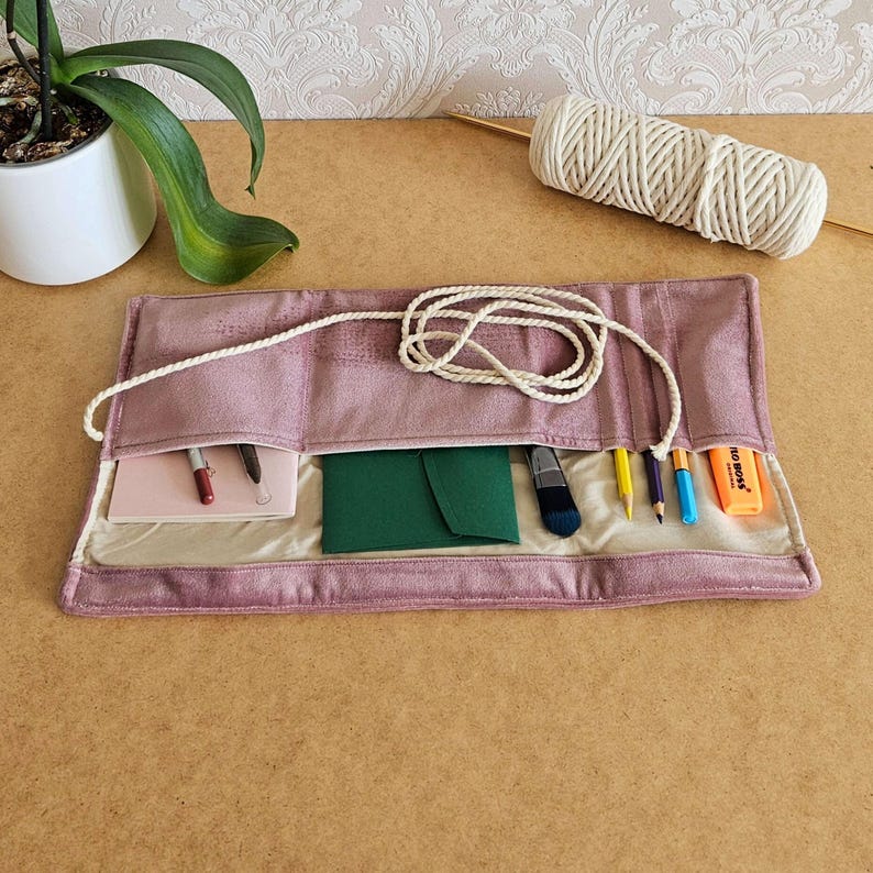 May include: A dusky pink roll-up organiser with multiple pockets, containing a notepad, pens, and stationery. A white cord secures the organiser. A spool of white cord and a plant are in the background.