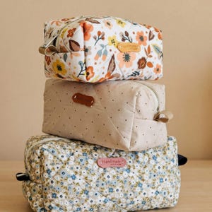 May include: Three stacked quilted cosmetic bags. The top bag features a white background with orange, yellow, and brown floral patterns. The middle bag is beige with small brown dots. The bottom bag has a blue floral pattern.