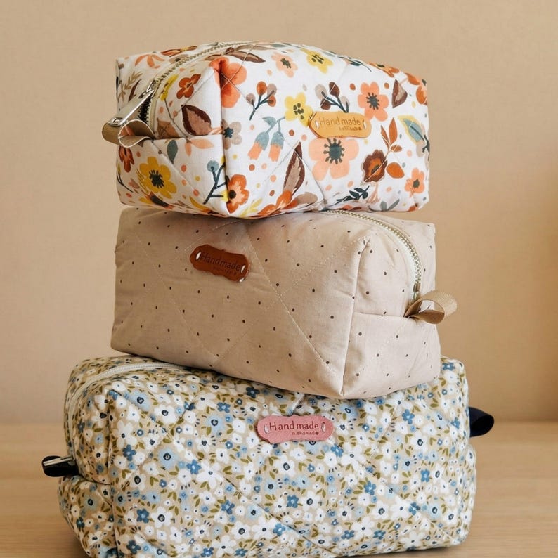May include: Three stacked quilted cosmetic bags. The top bag features a white background with orange, yellow, and brown floral patterns. The middle bag is beige with small brown dots. The bottom bag has a blue floral pattern.