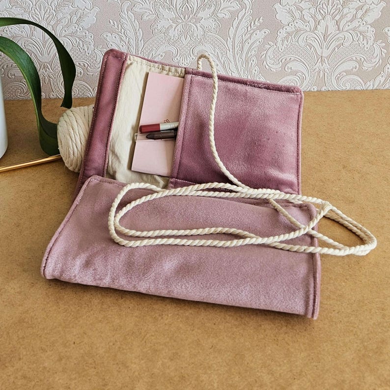 May include: A dusky pink velvet craft organiser, open to show a cream interior with pockets holding small notebooks and pens. A cream rope is attached for closure. A ball of yarn is visible on the side.