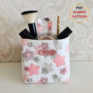 May include: A handmade fabric makeup bag with a star pattern in pink and gray, holding makeup brushes and cosmetics. The bag has a small handle and a "Handmade with Love" label. The image also includes text that says "PDF Sewing Pattern".