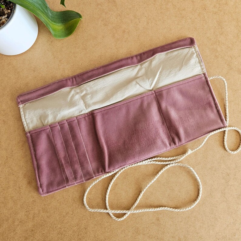 May include: A dusty rose leather roll-up case, open to reveal a cream-coloured interior and multiple pockets. A cream-coloured rope is attached. The case is on a light brown surface.