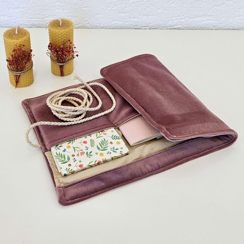 May include: A dusky pink velvet clutch, open to show a floral wallet and a small pink notebook. A white rope is coiled on top. Two yellow beeswax candles with dried flower decorations are in the background.